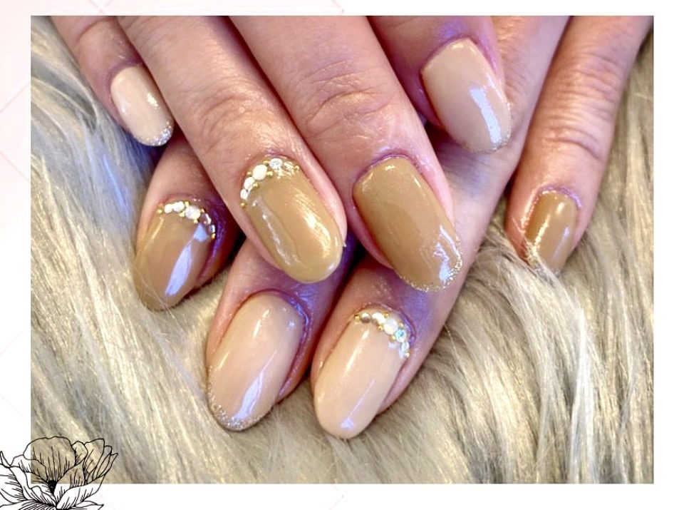 A close-up view of well-manicured hands with a mix of light beige and brown nail polish, decorated with small rhinestones along the tips.